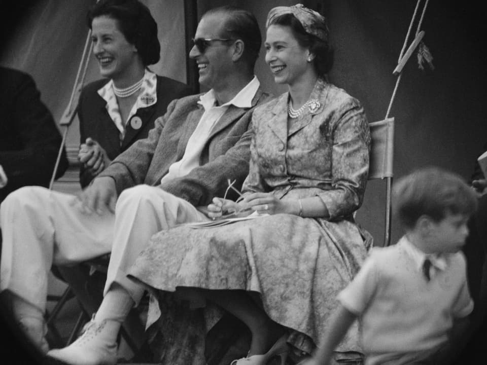Ein Mann mit Sonnenbrille und lockerem Anzug sitzt lachend nebend einer Frau im Kleid.