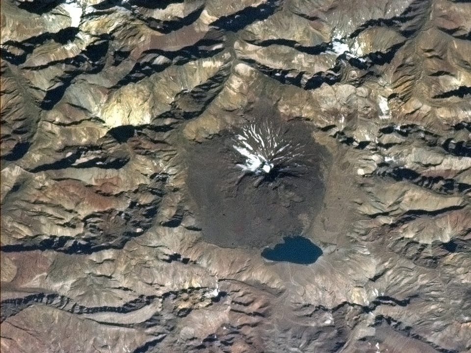 Blick aus dem Orbit auf einen rauchenden Vulkan in den Anden. An seinem Fuss liegt ein kleiner Kratersee.