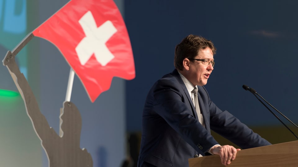 Albert Rösti an einem Rednerpult von der Seite. Am Rand ist eine Figur mit einer Schweizer-Fahne.