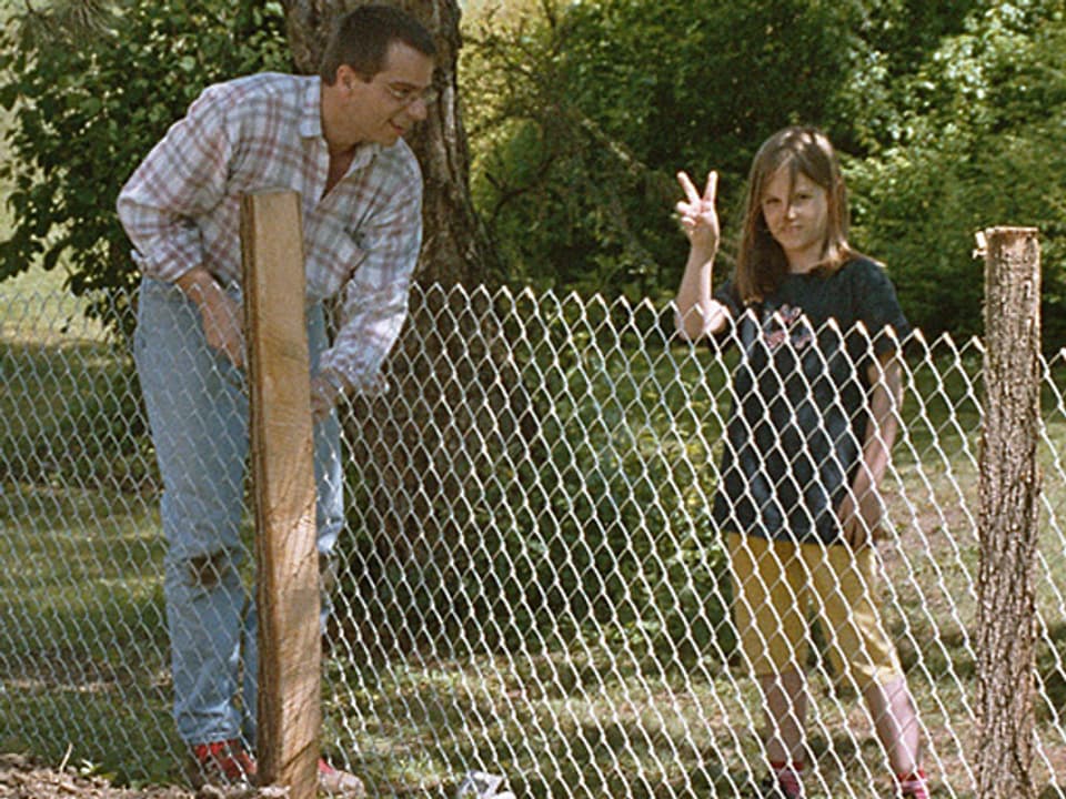 Ein Mann hält einen Holzpflock auf einer Wiese, daran ist ein Gitter befestigt. Daneben steht ein junges Mädchen und blickt in die Kamera.