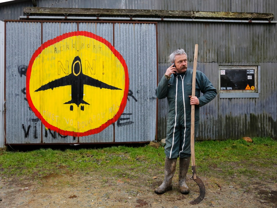 Ein Bauer mit einer Sichel und einem Handy am Ohr. Im Hintergrund ein Stoppschild mit einem Flugzeug drin auf eine Scheune gemalt.