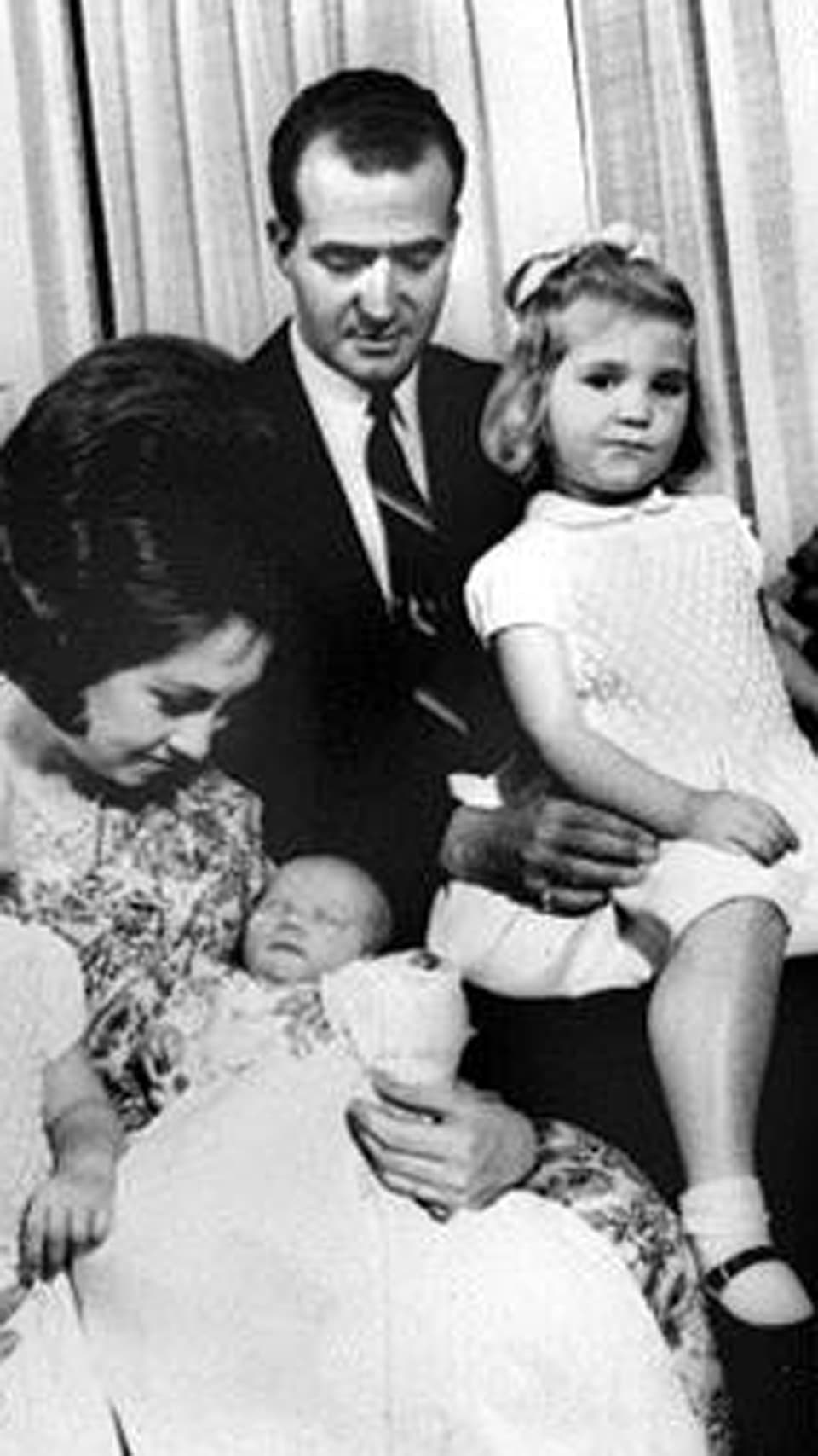 Familienfoto in schwarzweiss: Königin Sophia hält Baby Felipe auf dem Arm. Im Hintergrund sitzt seine Schwester in weissem Kleid auf der Schoss von König Juan Carlos.