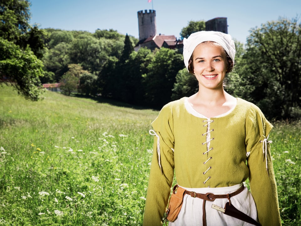 Junge Frau in mittelalterlicher Kleidung auf einer Wiese mit einer Burg im Hintergrund