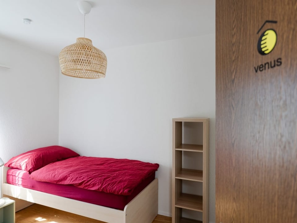 Ein Einzelzimmer mit einem 90er Bett und roter Bettwäsche. Die Türe mit dem Namenszeichen venus sieht man auch noch.
