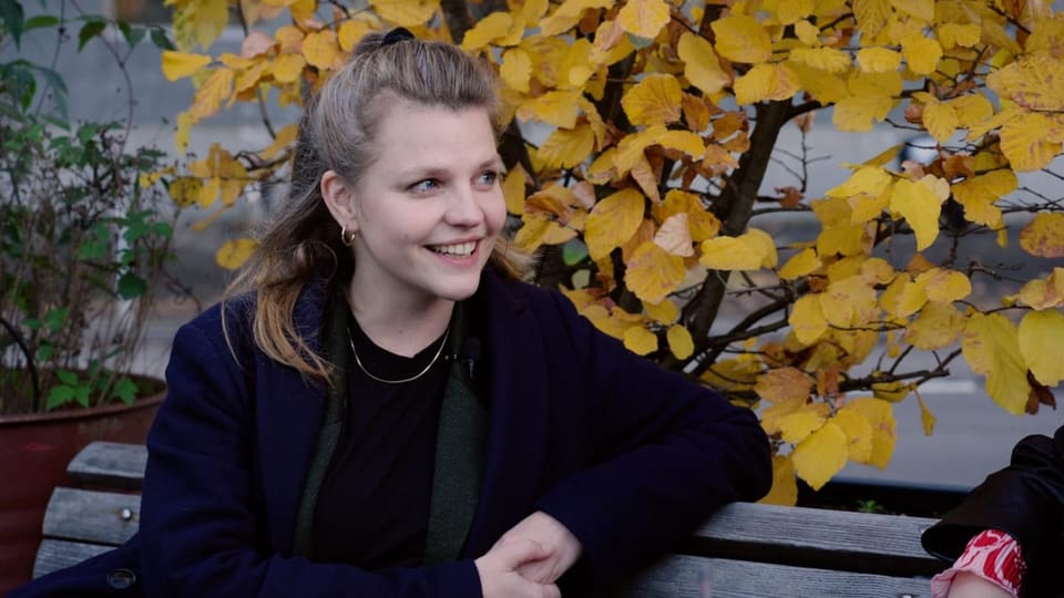 Die Sexualpädagogin Angie Walti sitzt auf einer Bank vor einem gelben Blätter-Strauch.