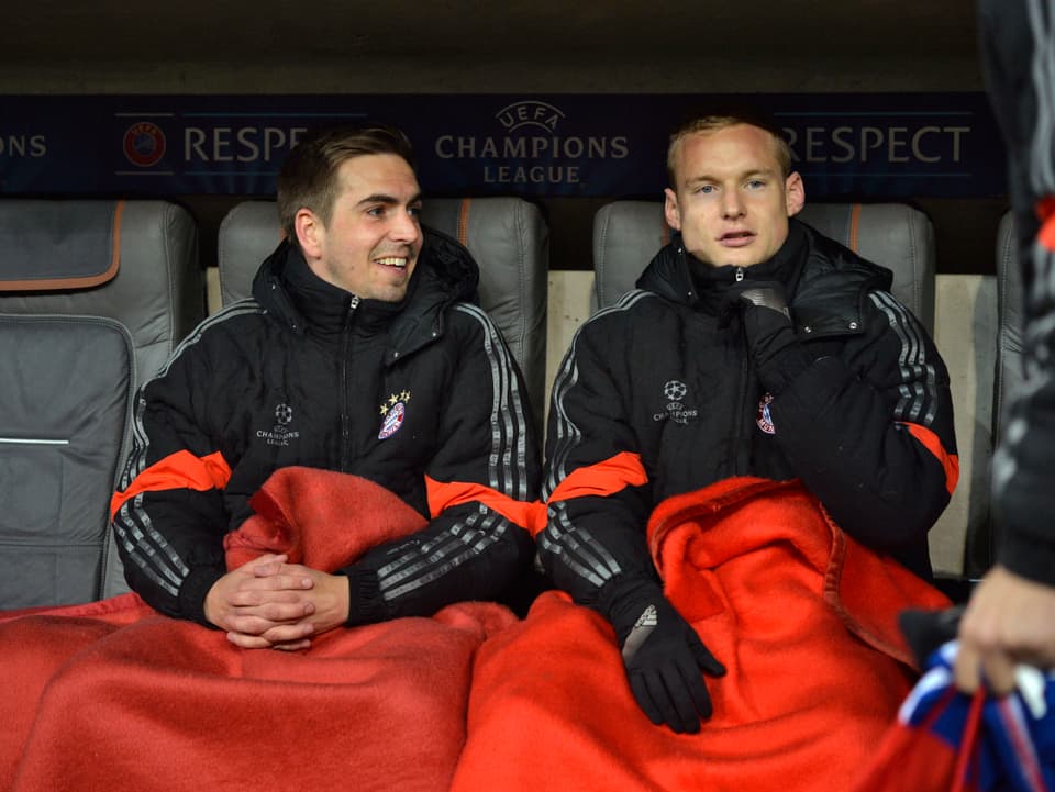 Philipp Lahm sitzt zusammen mit Bayern-Teamkollege Sebastian Rode auf der Ersatzbank, beide eingehüllt in rote Wolldecken.