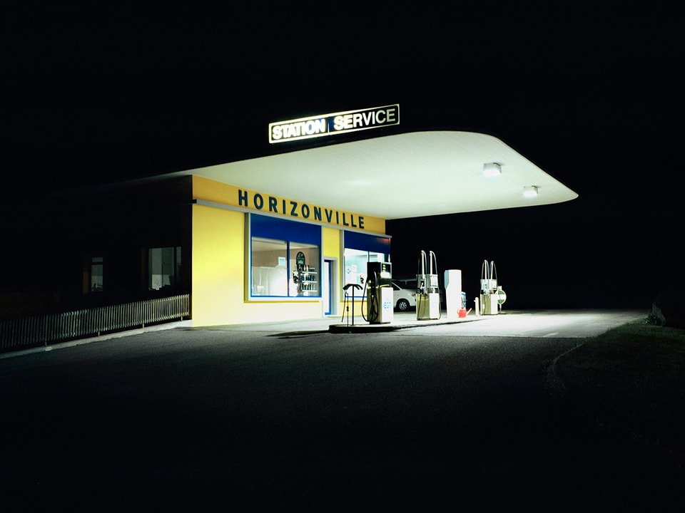 Eine hell beleuchtete, kleine Tankstelle mit zwei Zapfsäulen bei Nacht. An der Fassade steht "Horizonville".