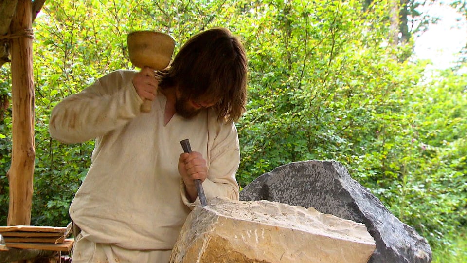 Mann in Jutehemd schlägt mit Holzhammer auf Meissel.