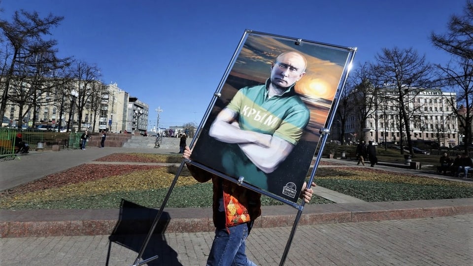 Ein Mensch stellt ein Plakat auf, das den russischen Präsidenten zeigt, als Illustration vor einem Sonnenaufgang.