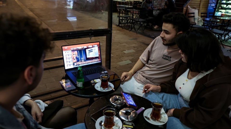 Menschen sitzen in einem Café um einen Laptop mit Newssendung