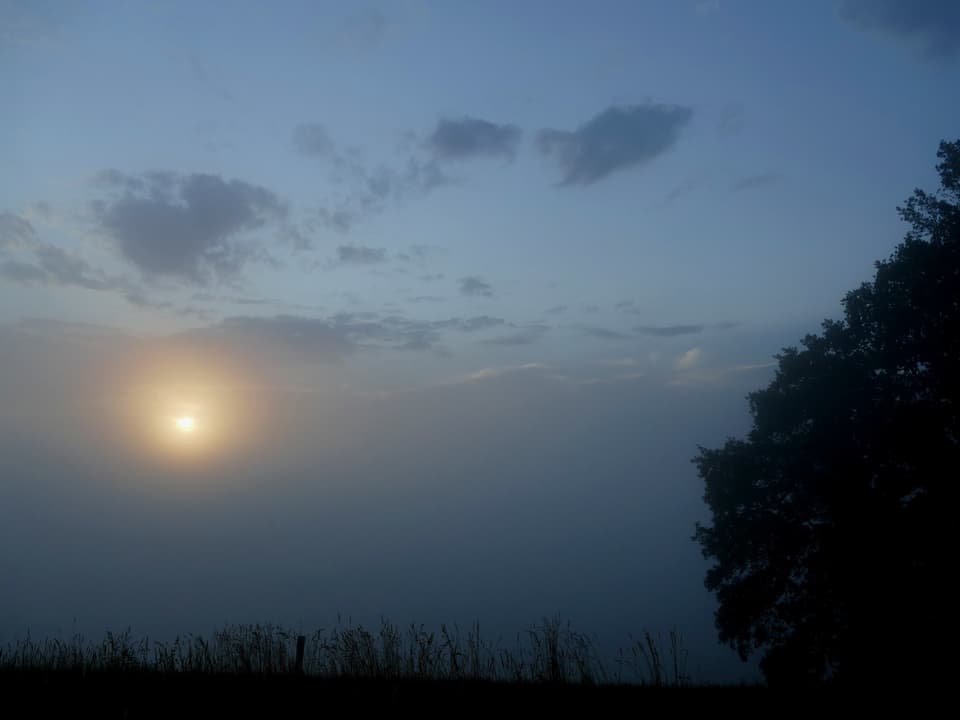 Ein dickes, graues Band liegt in der unteren Bildhälfte. Hinter dem Nebel geht die Sonne auf.