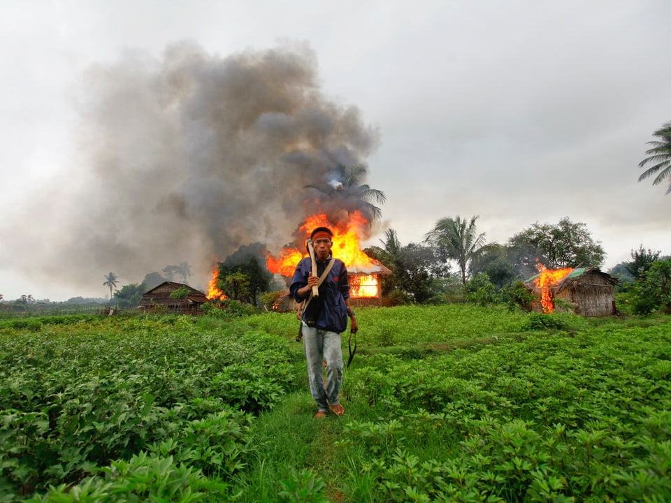 Militanter Buddhist mit Waffe, dahinter ein brennendes Haus.