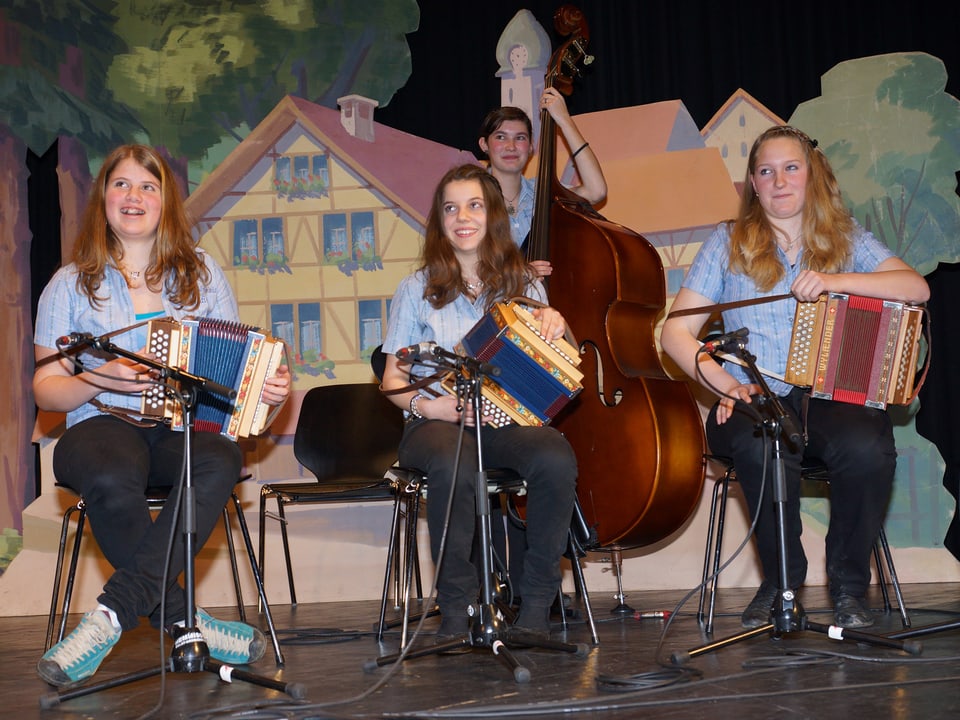 Vier junge Musikantinnen mit drei Schwyzerörgeli und einem Kontrabass sitzen und stehen lachend auf der Bühne.