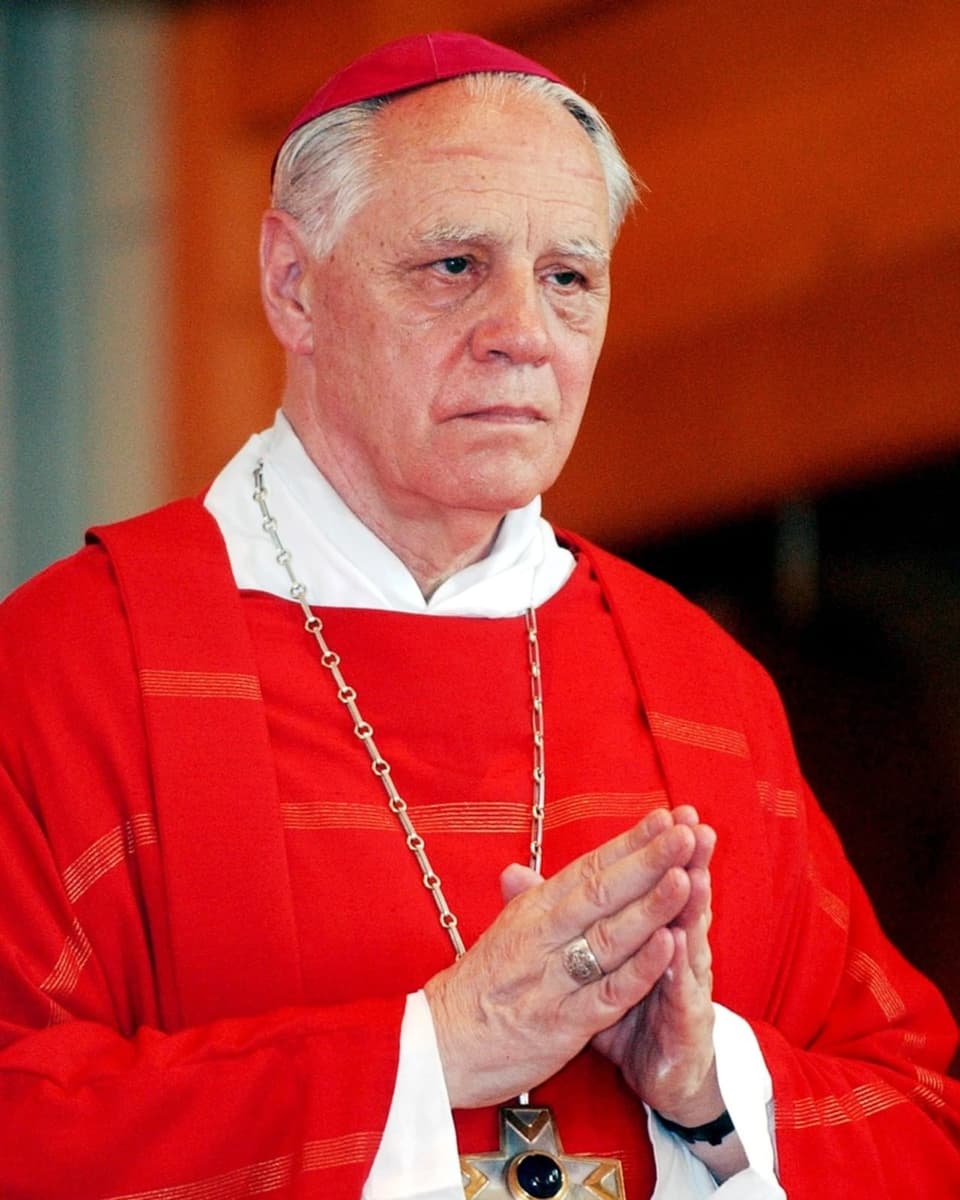 Älterer Mann in rotem Gewand, die Hände zum Gebet gefaltet. Er trägt eine Kette mit Kreuz.