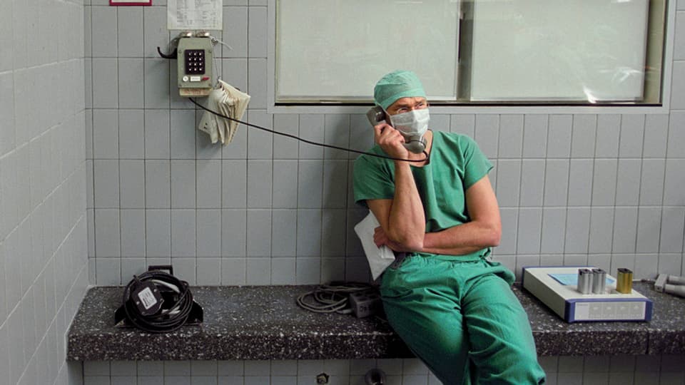 Mann mit grünem Arztkittel sitzt vor Operationssaal am Telefon. 