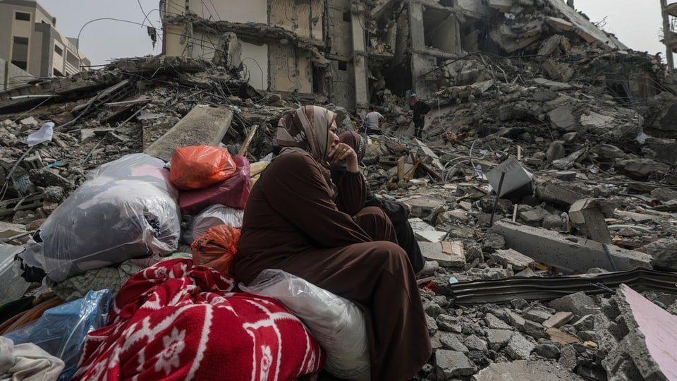Eine Frau sitzt mit Kleidersäcken und Decken vor einem zerstörtem Gebäude. 