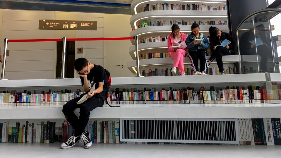 Drei junge Frauen sitzen nebeneinander in einer Bibliothek auf einer Bank und lesen.