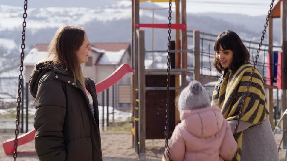 Moderatorin und Protagonisten stehen auf dem Spielplatz. Ein Kind sitzt auf der Schaukel.