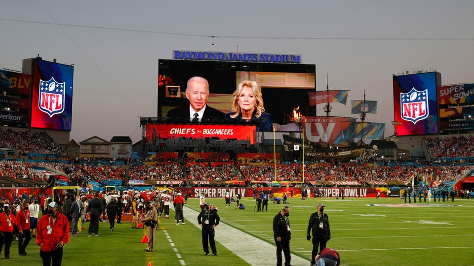 Ein Football-Stadion mit Menschen und im Hintergrund läuft eine Videobotschaft von Joe und Jill Biden. 
