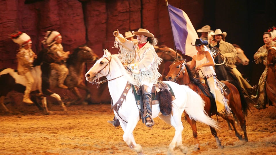 Ein Mann mit Cowboyhut reitet auf einem Pferd, hinter ihm weitere Reiter*innen.