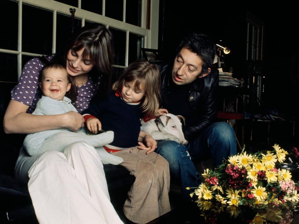 Foto aus den 70er Jahren, junge Familie mit zwei Töchtern, ein Baby und eine 5-jährige, sitzen auf dem Sofa und schauen zum Baby.