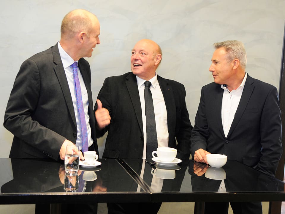 Die drei Männer führen an einem Tisch ein Gespräch.