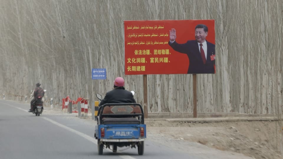 Eine Strasse auf der zwei motorisierte Fahrzeuge an einem grossen roten Schild mit Xi Jinping vorbei fahren. 