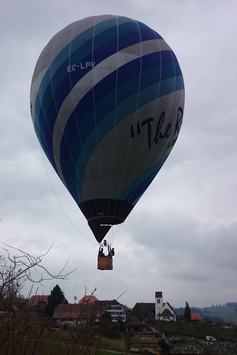 Ein Weiss-Blauer Ballon fliegt vor grauem Himmel. Im Hintergrund ist ein Dorf zu erkennen.