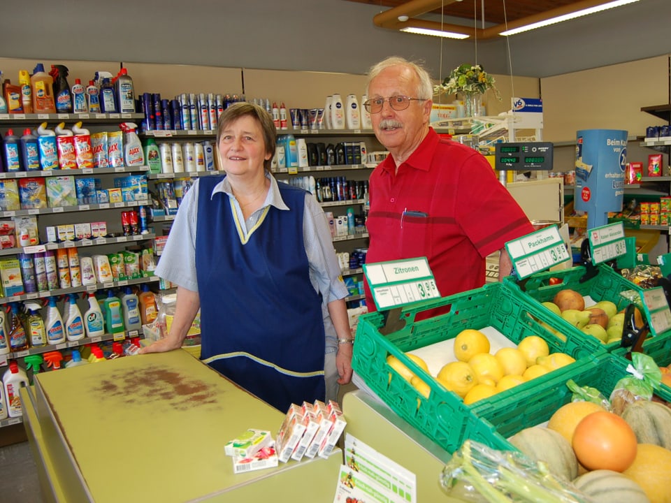 Die beiden Personen stehen hinter der Ladentheke und vor mit verschiedenen Produkten gefüllten Regalen.