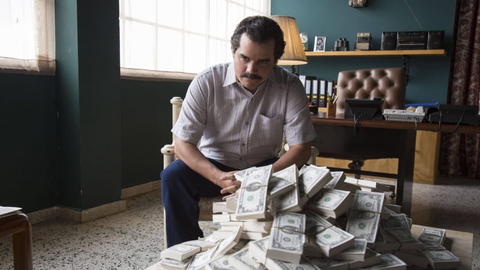 Pablo Escobar in der Fernsehserie sitzt vor einem Haufen Geldbeutel