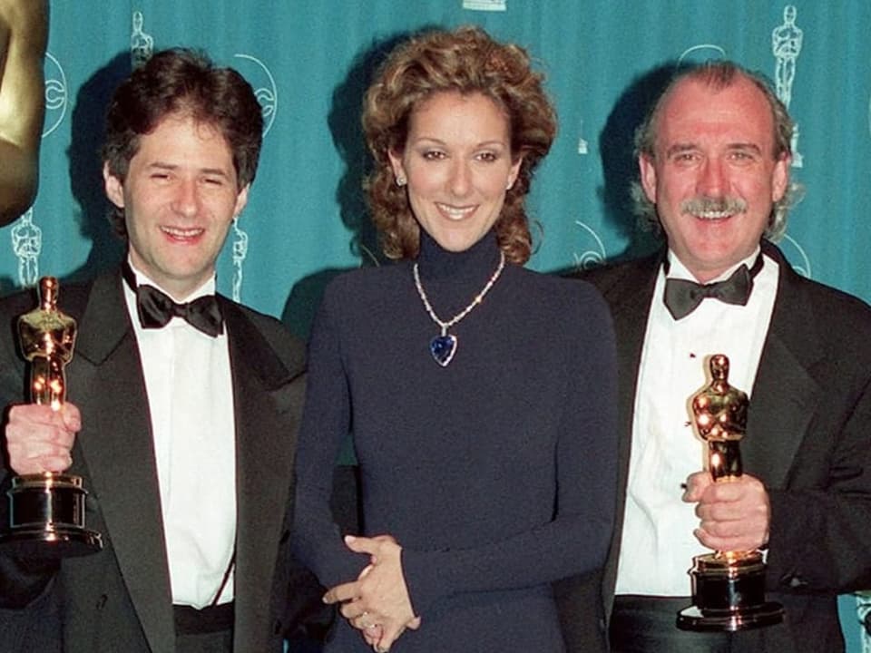 Céline Dion steht neben zwei Männern die je einen Oscar in der Hand halten.