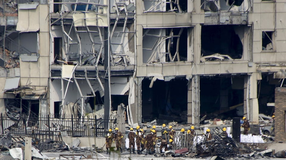 Zerstörtes Gebäude auf dem Hafengelände mit Rettungskräften in gelben Helmen davor