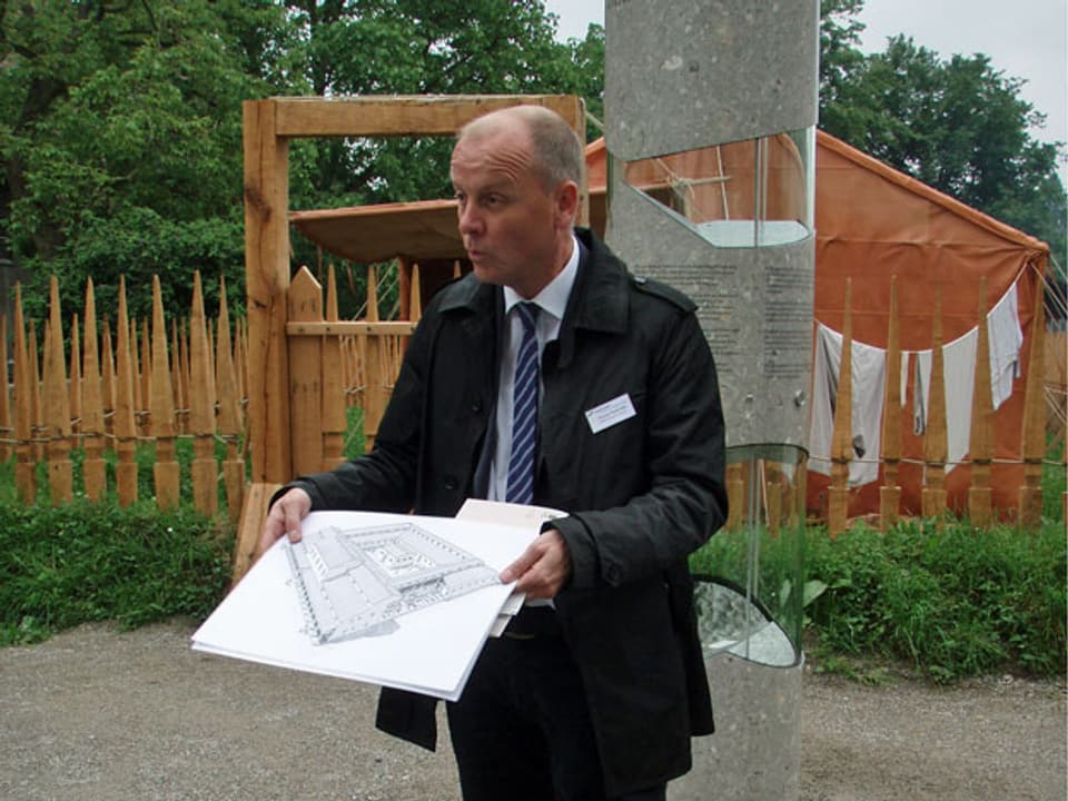 Thomas Pauli steht vor dem Lazarett-Eingang, in Schlips und Kravatte, und hält eine Zeichnung von einem Gebäudegrundriss in den Händen.