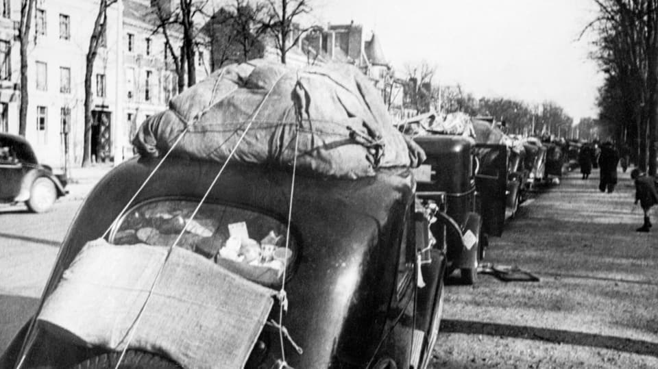 schwarzweiss-Foto alter Autos, eine ganze Kolonne davon, mit Bündeln auf Autodächer geschnürt, rechts gehen Menschen.