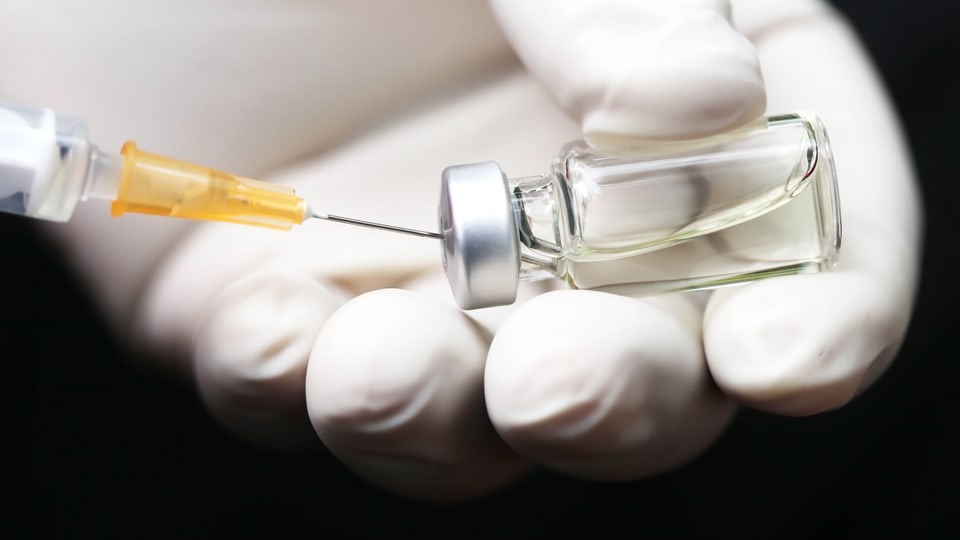 Es ist eine Nadel in einer Impfdose, welche von einer Hand mit Handschuh gehalten wird, abgebildet.