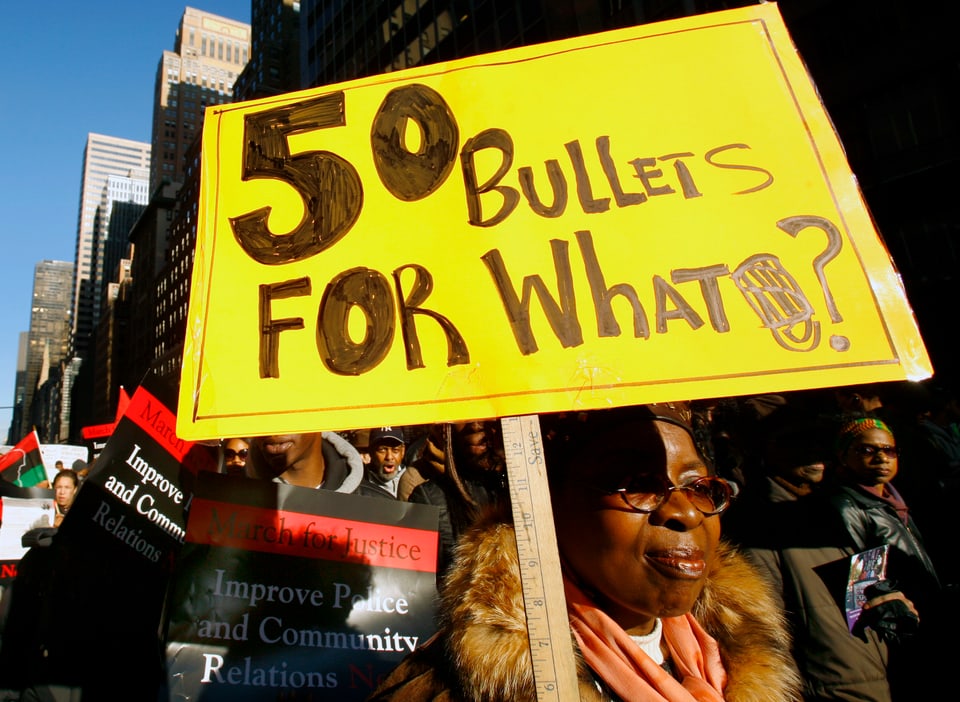 Eine Frau hält ein gelbes Schild mit der Aufschrift «50 Bullets for what?»