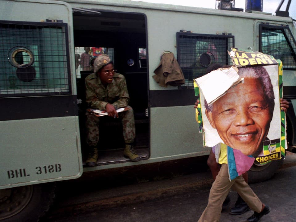 Ein Mann trägt ein zerissenes Plakat von Nelson Mandela an einem Polizeiwagen vorbei.