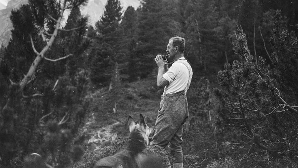 Hermann Langen steht mitten im Wald, ein Fenrglas halb zu den Augen gehoben und schaut angestrengt in die Ferne. Am unteren Bildrand schaut sein Schäferhund zu ihm auf.