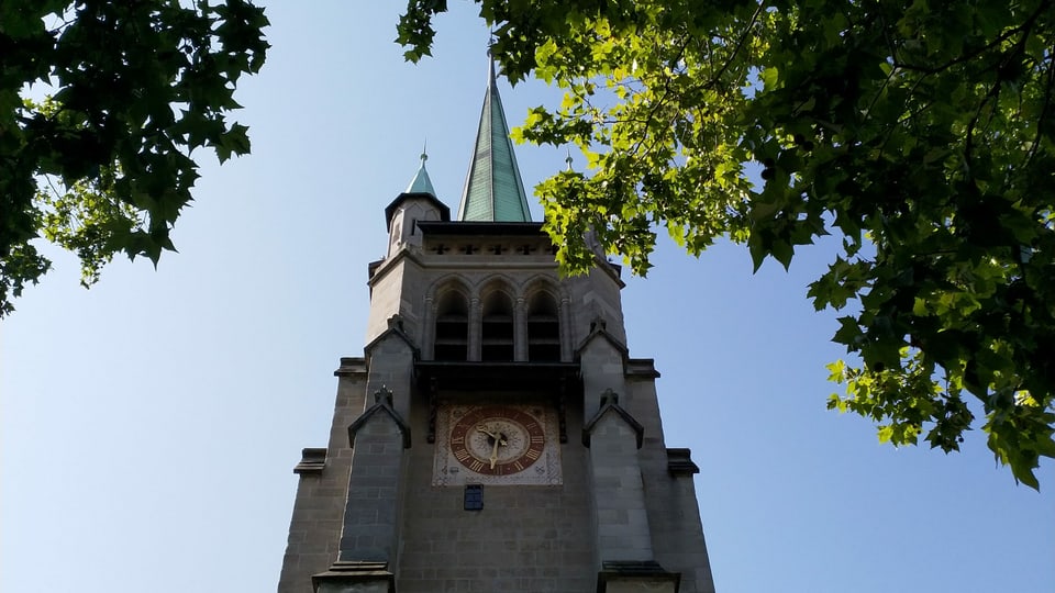 Durch Ahornblätter hindurch ist ein Kirchenturm mit rot-goldenem Ziffernblatt zu sehen.