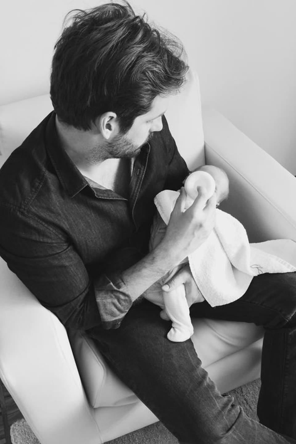 Schwarzweiss-Bild von Tomaso Trussardi auf einem Sessel sitzend und einem Baby den Schoppen gebend.