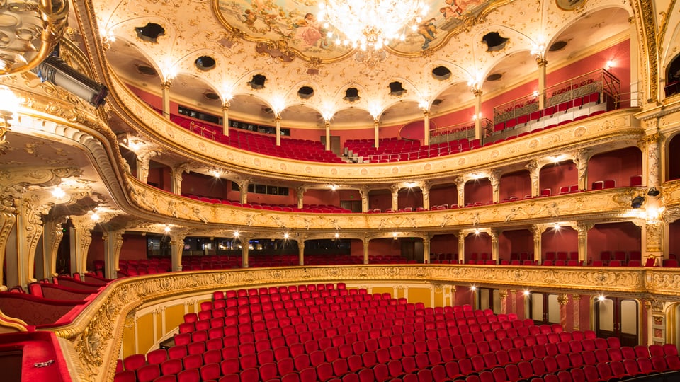 Blick in den Zuschauerraum des Opernhauses mit roten Sitzen und einem grossen Kronleuchter. 