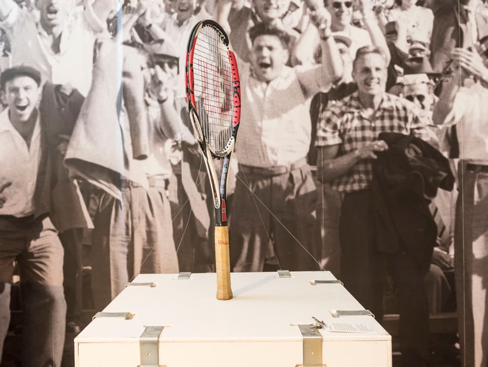 Tennisraket steht auf Ausstellungstisch, im Hintergrund sieht man jugendle Sportfans auf einem Foto.