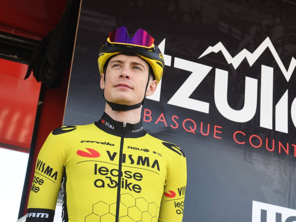 Radsportler in gelbem Trikot und violettem Helm steht vor einem Werbebanner.