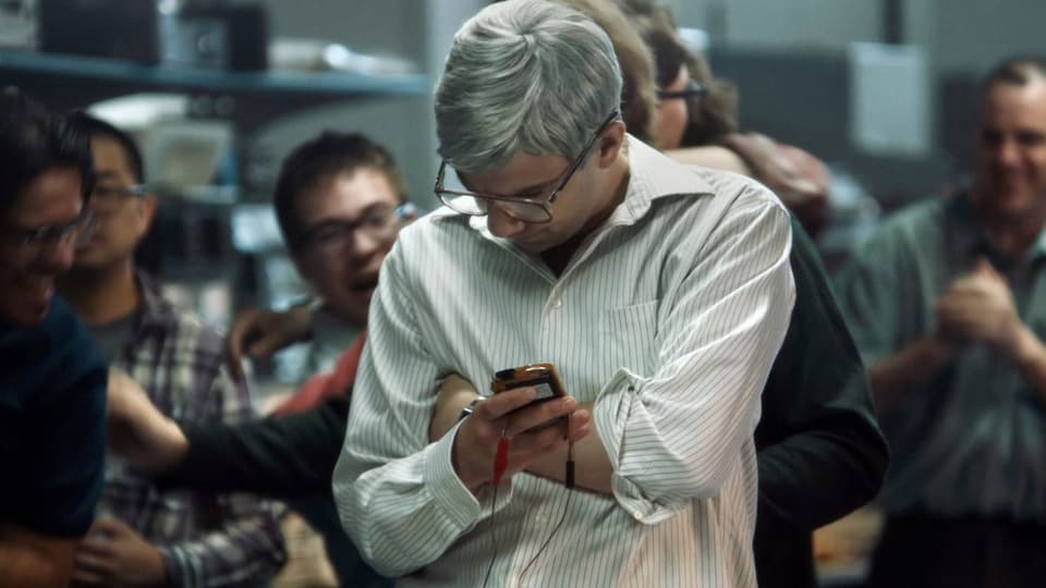 Ein Mann im Hemd blickt konzentriert auf ein Smartphone, im Hintergrund lachen mehrere Männer.