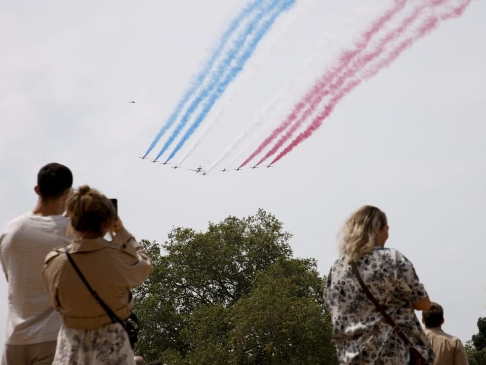 The planes emit smoke with British colors of blue, white and red.  Below, people watch the show. 