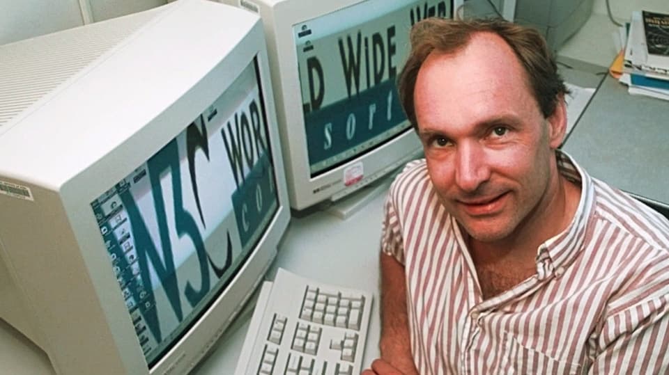 Mann Mitte 40. gestreiftes Hemd, sitzt, schaut auf. Hinter ihm zwei grosse Kastencomputer mit «world wide web» darauf.