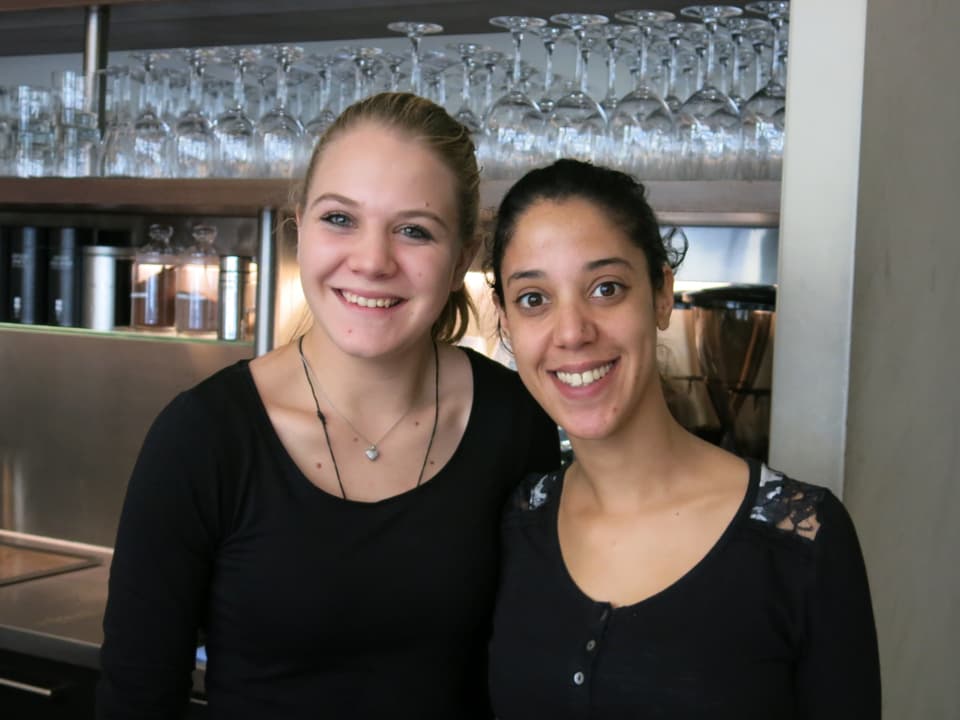 Zwei junge Frauen stehen vor einem Gläser-Buffet und lachen in die Kamera.