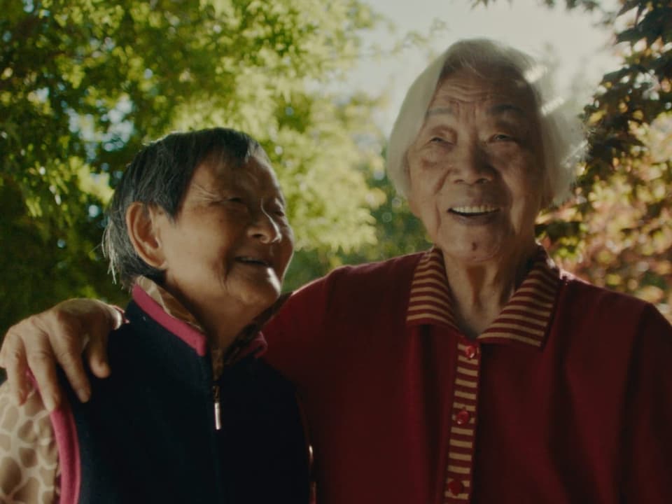 Zwei alte Frauen liegen sich in den Armen und lächeln. Im Hintergrund Bäume.
