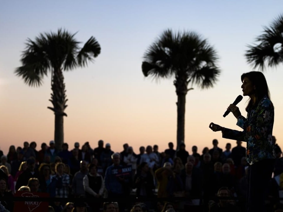 Nikki Haley, Wählerinnen und Wähler sowie Palmen erscheinen im Gegenlicht. Im Hintergrund: der Himmel im Dämmerlicht.