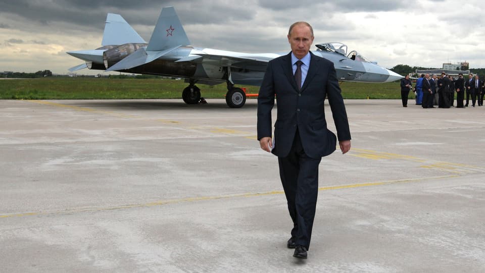 Putin auf einem Militärstützpunkt, hinter ihm ein stehendes Kampfflugzeug und eine Gruppe Leute.